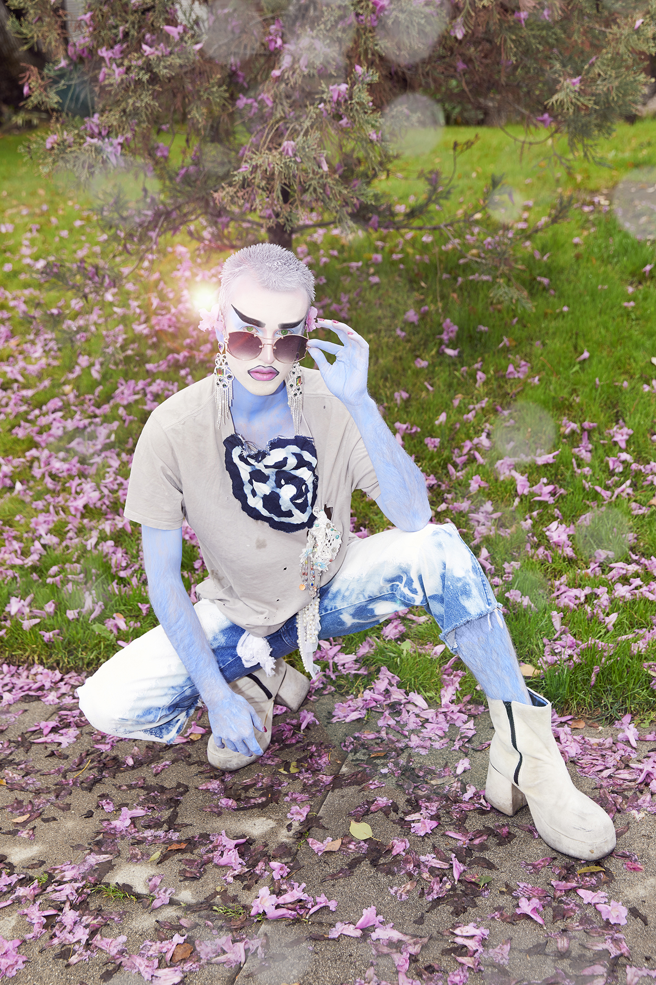 Jesse in blue makeup crouched on a sidewalk covered in pink flowers